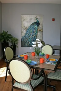 A dining table set for four with colorful dishes and mugs, a vase of white flowers, green upholstered chairs, and a large peacock painting on the gray wall behind. A dining table set for four with colorful dishes and mugs, a vase of white flowers, green upholstered chairs, and a large peacock painting on the gray wall behind.